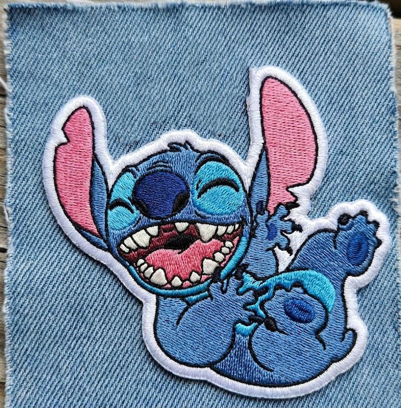 May include: Embroidered patch of the character Stitch from Lilo & Stitch. The patch shows Stitch in blue, with pink inner ears and mouth, and a white outline. The patch is set on a denim background.