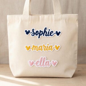 Może przedstawiać: Kremowa torba typu tote z trzema haftowanymi naszywkami z imionami: "Sophie" w kolorze granatowym, "Maria" w kolorze ż&oacute;łtym i "Ella" w kolorze r&oacute;żowym. Każde imię jest stylizowane czcionką inspirowaną Disneyem i uszami Myszki Miki.