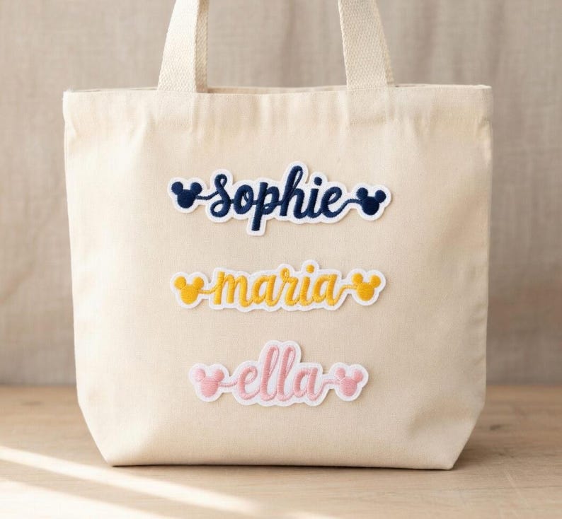 Może przedstawiać: Kremowa torba typu tote z trzema haftowanymi naszywkami z imionami: "Sophie" w kolorze granatowym, "Maria" w kolorze ż&oacute;łtym i "Ella" w kolorze r&oacute;żowym. Każde imię jest stylizowane czcionką inspirowaną Disneyem i uszami Myszki Miki.