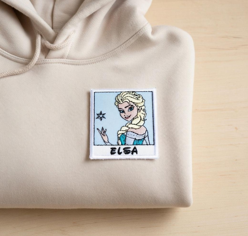 May include: A beige hoodie with a square patch featuring an embroidered image of Elsa from Frozen. Elsa is depicted with blonde hair and a blue dress, with a snowflake and the name "ELSA" below. The hoodie is made of a soft material.