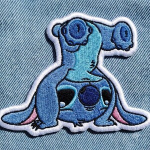 May include: Embroidered patch of a blue cartoon character with large ears and eyes, outlined in white. The character is in a playful pose, with pink accents on the ears and feet. The patch is designed for clothing or accessories.