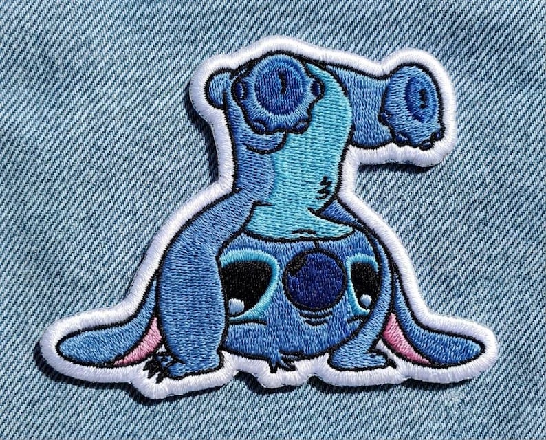 May include: Embroidered patch of a blue cartoon character with large ears and eyes, outlined in white. The character is in a playful pose, with pink accents on the ears and feet. The patch is designed for clothing or accessories.