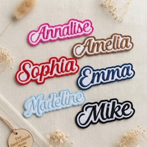 Może przedstawiać: Haftowane naszywki z imionami w różnych kolorach, w tym różowym, brązowym, czerwonym, niebieskim, jasnoniebieskim i czarnym. Każda naszywka zawiera inne imię w kursywie z kontrastującą obwódką. Imiona to Annalise, Amelia, Sophia, Emma, Madeline i Mike.