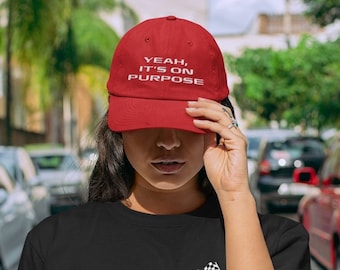 Puede incluir: Una mujer con una camiseta negra con un diseño de bandera a cuadros blanca y una gorra de béisbol roja que dice "Yeah, it's on purpose".