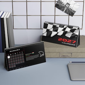 May include: A 2027 desk calendar featuring a black and white checkered flag design on the cover. The open page displays the month of August with a grid, a small map of the Budapest, Hungary race track, and descriptive text in English.