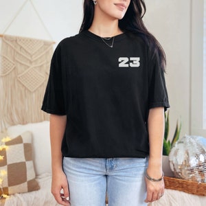 May include: A black t-shirt with the number "23" printed in white on the front. The shirt is being worn by a person with long dark hair.