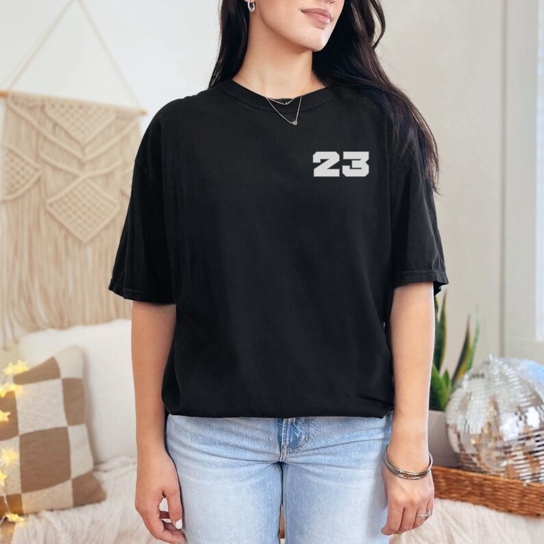 May include: A black t-shirt with the number "23" printed in white on the front. The shirt is being worn by a person with long dark hair.