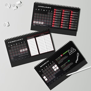 May include: Three black spiral-bound desk calendars for the 2027 racing season. The pages display January, February, and March with grids, race schedules, driver prediction charts, and track maps. A white pen rests near the March calendar.