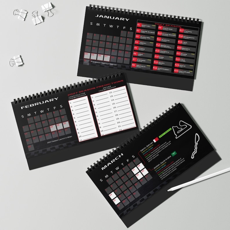 May include: Three black spiral-bound desk calendars for the 2027 racing season. The pages display January, February, and March with grids, race schedules, driver prediction charts, and track maps. A white pen rests near the March calendar.