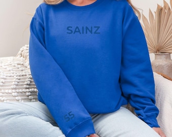 May include: A royal blue sweatshirt with the word "SAINZ" printed on the front in a dark blue color. The sweatshirt has a relaxed fit and a crew neck. The number "55" is printed on the left sleeve.