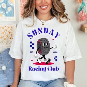 以下が含まれることがあります： 白いTシャツに、顔が付いたタイヤの漫画と「SUNDAY Racing Club」の文字、タイヤの下に「EST. 1950」の文字が書かれた青と白のグラフィックがプリントされています。