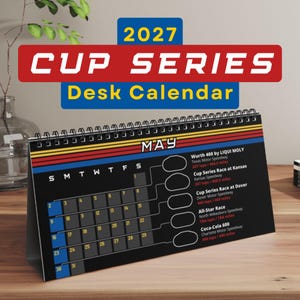 May include: A 2027 Cup Series desk calendar with a black background, spiral binding, and a May monthly grid. The design features red, yellow, and blue horizontal stripes, with race event details listed on the right side for the month of May.