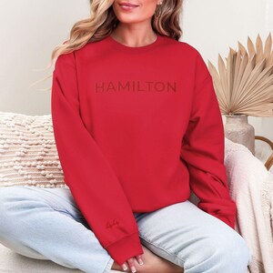 Op de afbeelding: Rood sweatshirt met het woord "HAMILTON" geborduurd in rood op de voorkant. Het sweatshirt heeft een ronde hals en lange mouwen.