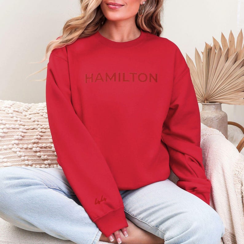 Op de afbeelding: Rood sweatshirt met het woord "HAMILTON" geborduurd in rood op de voorkant. Het sweatshirt heeft een ronde hals en lange mouwen.