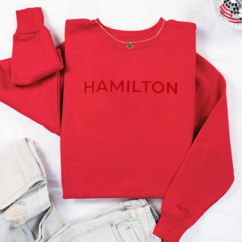 Op de afbeelding: Rood sweatshirt met het woord "HAMILTON" in wit op de voorkant. Het sweatshirt heeft lange mouwen en een ronde hals.