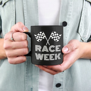 Op de afbeelding: Zwarte koffiemok met een wit ontwerp van twee gekruiste geblokte vlaggen met de tekst "RACE WEEK" eronder.