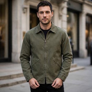 May include: An olive green zip-up jacket with a classic collar, long sleeves, and two slanted front pockets. The jacket is worn over a plain black shirt, featuring a central metal zipper closure and a structured, straight-cut silhouette.