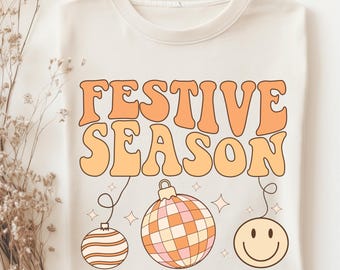 May include: A white t-shirt with the text "FESTIVE SEASON" in orange and yellow letters. The text is in a retro font style. There are three ornaments hanging from the text: a striped ornament, a checkered ornament, and a smiley face ornament.