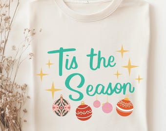 May include: A white t-shirt with the text "Tis the Season" in teal green lettering and gold stars. The shirt also features a row of five colorful Christmas ornaments.