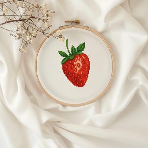 May include: A cross-stitch embroidery of a red strawberry with green leaves and stem, set in a wooden embroidery hoop. The strawberry is centered on a white fabric background. The background is a white, silky fabric.