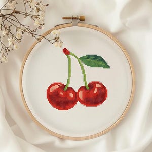 May include: Cross-stitch embroidery of two red cherries with green stems and a leaf, set against a white background within a wooden embroidery hoop. The cherries are rendered in a pixelated style.