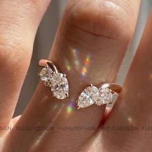 Könnte beinhalten: Ein offener Ring aus Roségold mit klaren, funkelnden Edelsteinen. Der Ring hat ein einzigartiges Design mit zwei separaten Abschnitten, die sich um den Finger legen, jeder mit mehreren Steinen besetzt.