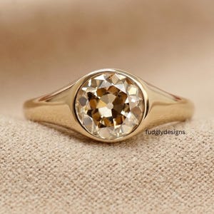 May include: A gold ring featuring a round, faceted champagne-colored gemstone set in a smooth, polished bezel setting. The band is solid gold with a tapered design that widens toward the center stone. The text fudglydesigns is visible.