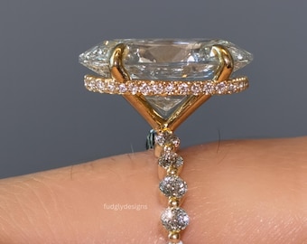 Puede incluir: Un anillo de compromiso de oro con una piedra preciosa grande y transparente de corte rectangular. El anillo tiene un halo de piedras más pequeñas alrededor de la piedra principal y piedras adicionales a lo largo de la banda. El anillo está sobre un fondo neutro.