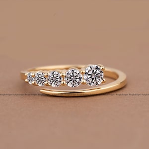 Puede incluir: Un anillo abierto de oro con cinco piedras redondas transparentes dispuestas en una secuencia de tamaño graduado a lo largo de la curva superior. La banda es de oro pulido y las piedras están engastadas en fila, creando un punto focal brillante.