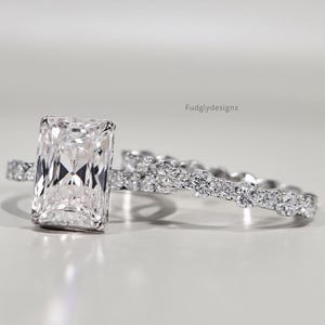 May include: A radiant-cut diamond engagement ring and matching band. The engagement ring has a large, rectangular diamond on a delicate band. The matching band is decorated with smaller diamonds. The rings are set against a soft, neutral background.