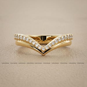 Pode incluir: Dois anéis dourados, um com pequenas pedras transparentes, o outro em forma de V. Ambos em fundo neutro. O nome da marca "FUDGLYDESIGN" está impresso na parte inferior. Anéis de noivado ou casamento.