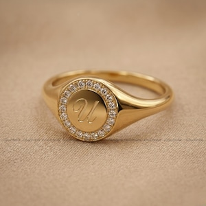 Op de afbeelding: Een gouden zegelring met een ronde voorkant. De voorkant is omcirkeld door kleine, heldere stenen en heeft de cursieve letter "U". De ring heeft een gepolijste, gladde afwerking.