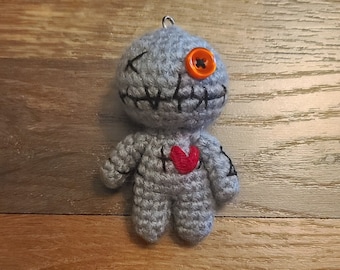 May include: A gray crocheted doll with a stitched-on mouth and one orange button eye. The doll has a red heart stitched on its chest.