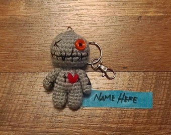 May include: A gray crocheted voodoo doll keychain with a red heart, black stitching, and an orange button eye. The keychain has a silver metal ring and a blue tag that says "NAME HERE".