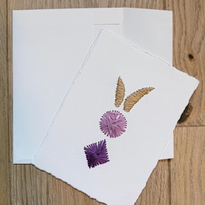 May include: A white greeting card with deckled edges featuring hand-stitched embroidery. The design includes two tan thread ears, a circular light purple thread motif, and a dark purple thread diamond shape, accompanied by a plain white envelope.