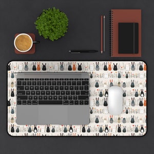 May include: A desk mat featuring a repeating pattern of cartoon bunnies in various colors and striped shirts, set against a cream background with floral accents. A laptop, mouse, and keyboard are on the mat, along with office supplies.