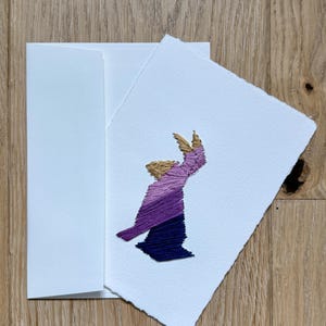May include: A white greeting card with a deckled edge featuring an embroidered figure in a gradient of purple and blue threads, accented with gold thread at the top. The card is paired with a plain white envelope.