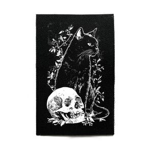 Puede incluir: Parche negro con una ilustración blanca de un gato negro sentado sobre una calavera, rodeado de follaje. El gato está de perfil. El fondo es negro.