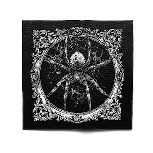 May include: A square black fabric patch featuring a white screen-printed illustration of a spider centred within an ornate, circular Victorian-style frame. The spider is depicted resting on a web, with intricate filigree details on the border.
