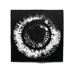 May include: A square, black patch with a white iris design. The iris has a dark central circle, radiating lines, and a textured outer ring. The design is printed on black fabric, creating a high-contrast visual effect.