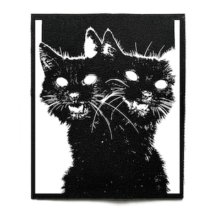 Puede incluir: Parche en blanco y negro con un gato de dos cabezas con ojos blancos brillantes y bocas abiertas. El pelaje del gato es detallado y el parche tiene un borde blanco. El diseño es una ilustración gráfica audaz.
