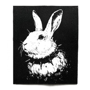 Pode incluir: Patch em preto e branco com um coelho com uma coleira de espinhos. O coelho é retratado em detalhe, em perfil. O fundo é preto sólido, criando um forte contraste com o coelho branco.