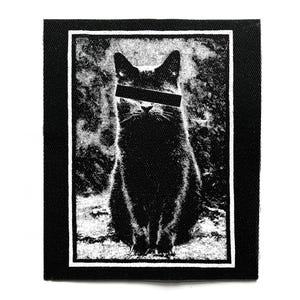 Puede incluir: Parche en blanco y negro con un gato estilizado con una barra negra sobre los ojos. El gato está centrado dentro de un rectángulo con borde blanco, sobre un fondo texturizado. El parche tiene un borde negro.