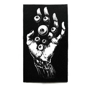 Puede incluir: Parche en blanco y negro con una ilustración detallada de una mano sosteniendo múltiples globos oculares. El diseño tiene un tema gótico o de terror, con un alto contraste entre el fondo negro y la mano y los globos oculares blancos.