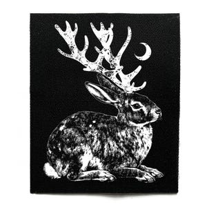 Könnte beinhalten: Schwarz-weißer Aufnäher mit einem Hasen mit Hirschgeweih und einem Halbmond. Die Illustration hat ein detailliertes, strukturiertes Aussehen. Der Hase sitzt, die Ohren aufgestellt. Das Geweih ist weiß und der Hintergrund ist einfarbig schwarz.