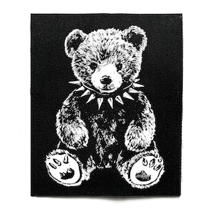 May include: Black and white patch depicting a teddy bear wearing a spiked collar. The bear is seated, with its paws clearly visible. The patch has a black background and a white border.