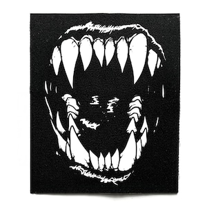 May include: Black fabric patch with a white illustration of a snarling mouth and sharp teeth. The design is a bold, graphic representation of a ferocious animal or creature, suitable for clothing or accessories.