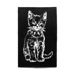 Peut inclure: Écusson noir et blanc représentant un chat portant un collier clouté. Le chat est assis, la queue enroulée. L'écusson est rectangulaire, avec un fond noir.