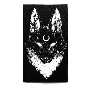 May include: A black rectangular patch featuring a white illustration of a wolf's head. The wolf has a crescent moon above its eyes. The design is detailed with textured shading, creating a striking contrast against the black background.