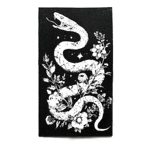 Op de afbeelding: Een zwart rechthoekig embleem met een witte illustratie van een slang verweven met bloemen en bladeren. De slang heeft zijn tong uit, en het ontwerp bevat sterren.